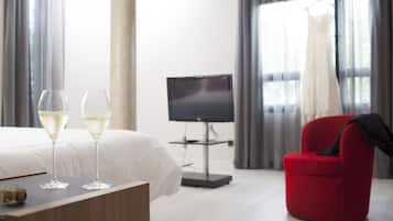 A hotel room with a bed, two glasses of white wine, a TV, and a red chair.