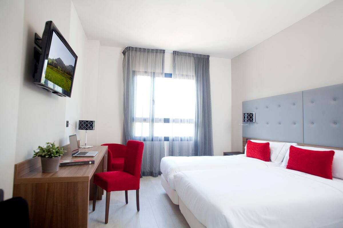 A hotel room with a bed, a desk with a laptop, a chair, a TV mounted on the wall, and a window with curtains.