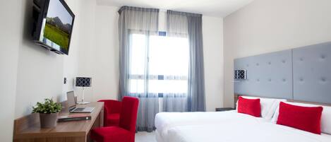A hotel room with a bed, a desk with a laptop, a chair, a TV mounted on the wall, and a window with curtains.