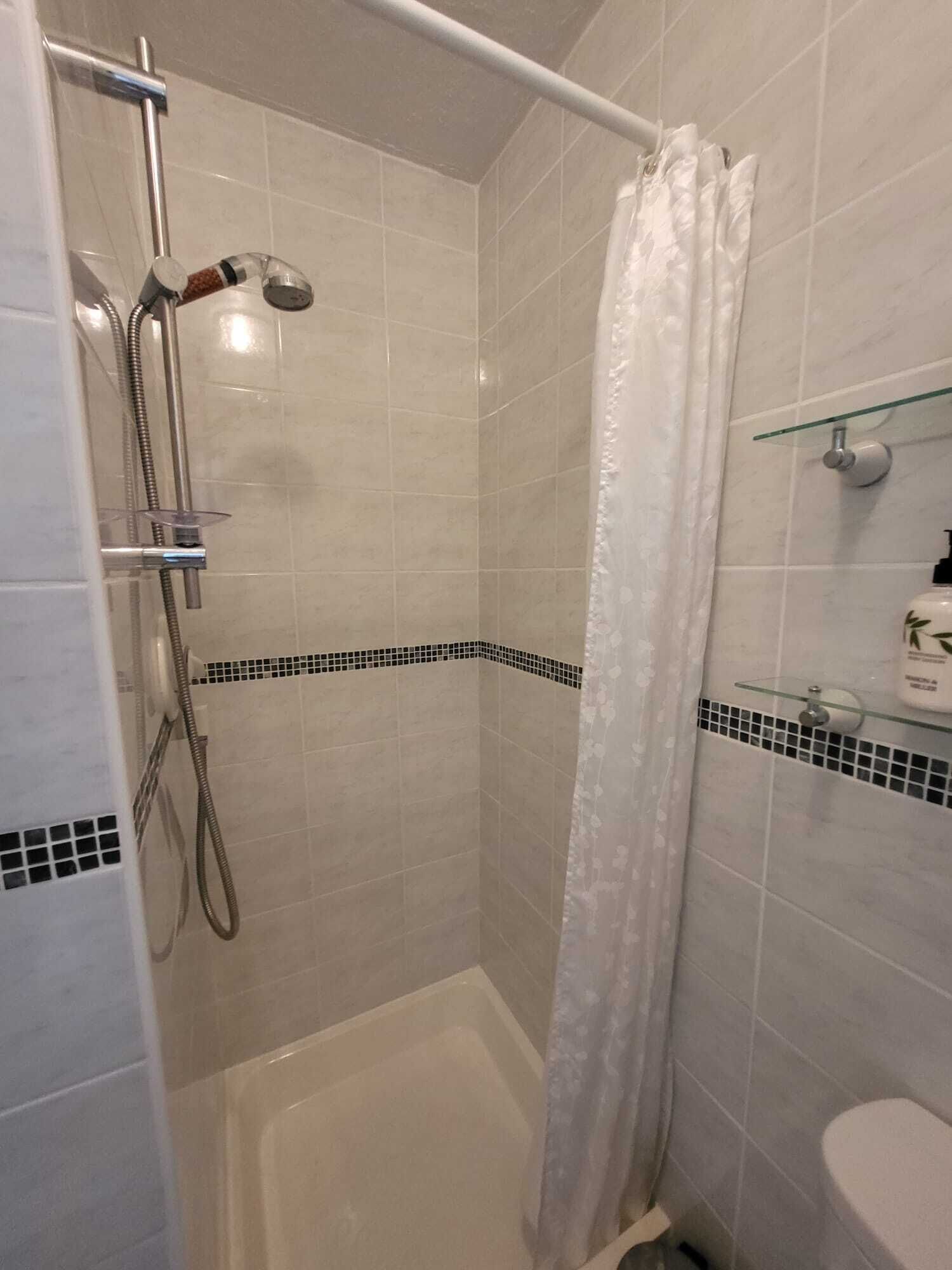A shower area with a glass door, a handheld showerhead, and a glass shelf with toiletries.