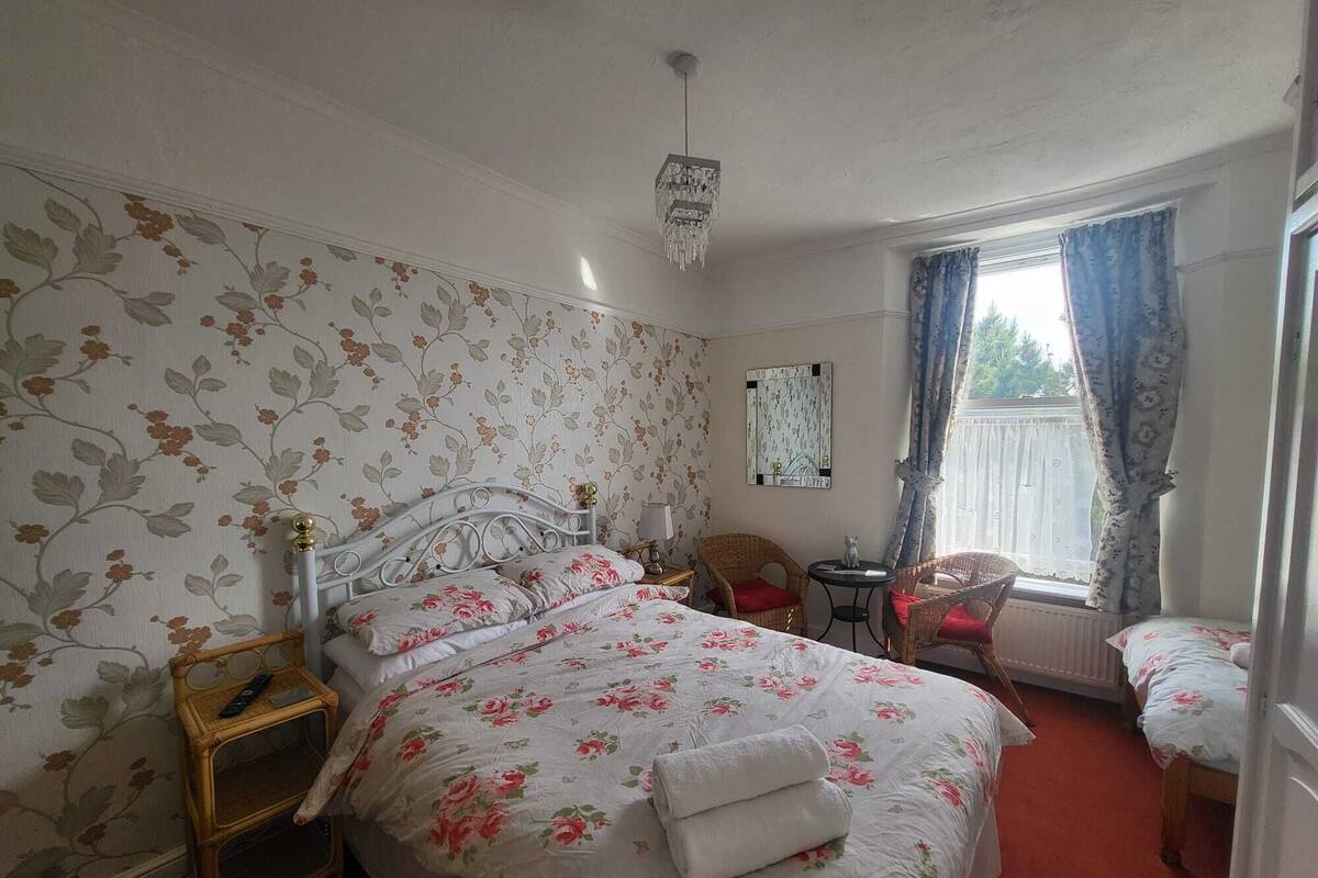 A bedroom with floral wallpaper, a bed with a white floral bedspread, a bedside table with a lamp, and a window with curtains.