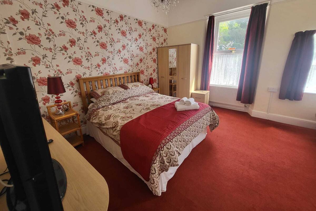 A bedroom with a floral wallpaper, a wooden bed, a red carpet, a chandelier, and a window with curtains.