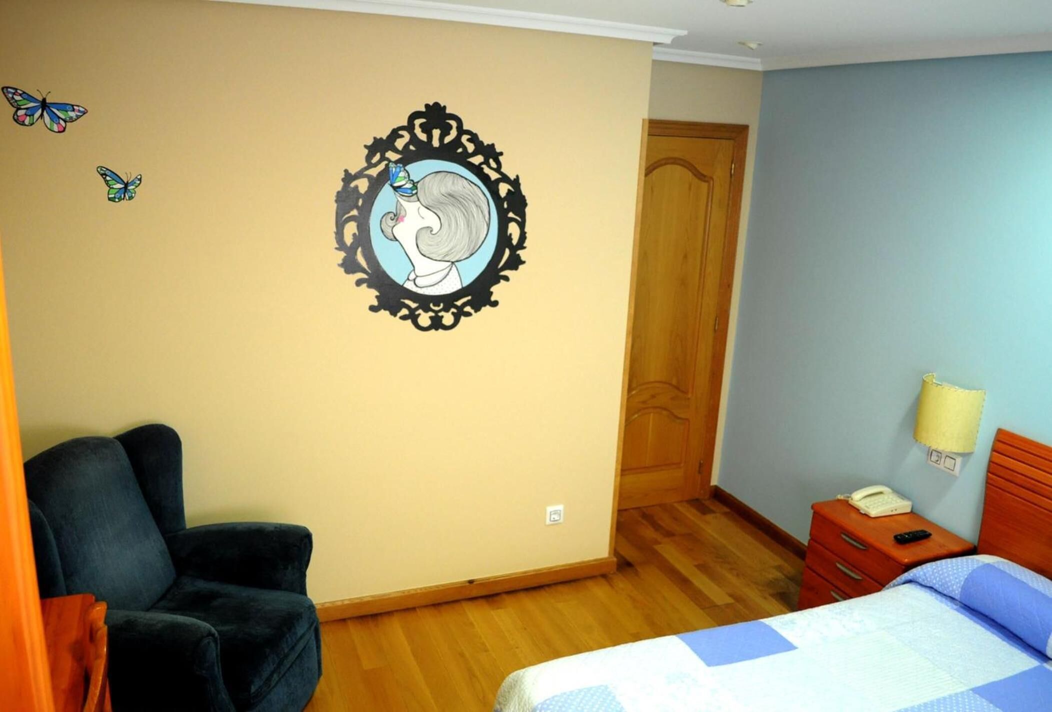 Une chambre à coucher comprenant un lit, une table de chevet, une chaise, une porte en bois et un mur orné d’un motif autocollant représentant un papillon.