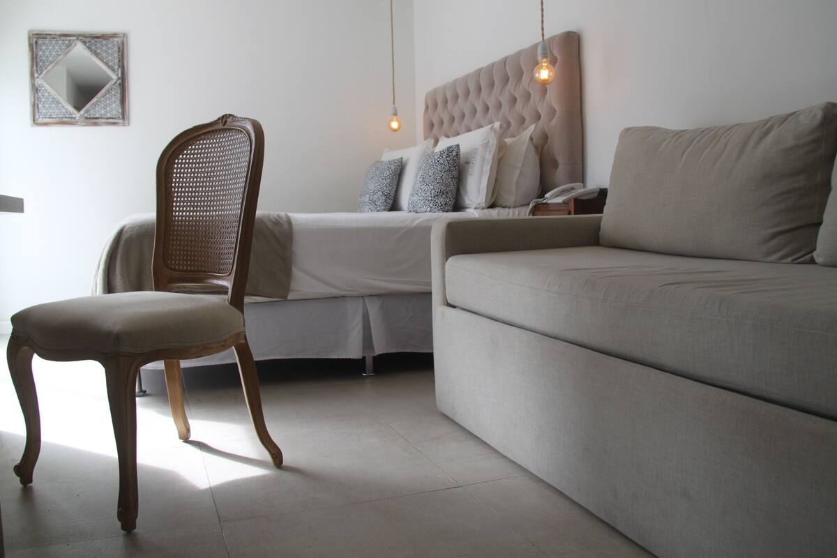 A bedroom with a bed, a wooden chair, a sofa, and a nightstand with a lamp.
