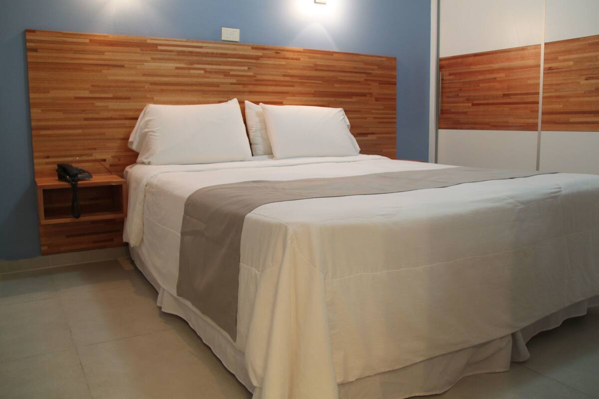 A neatly made bed with white linens and a beige throw blanket, a wooden headboard, and a bedside table with a phone.