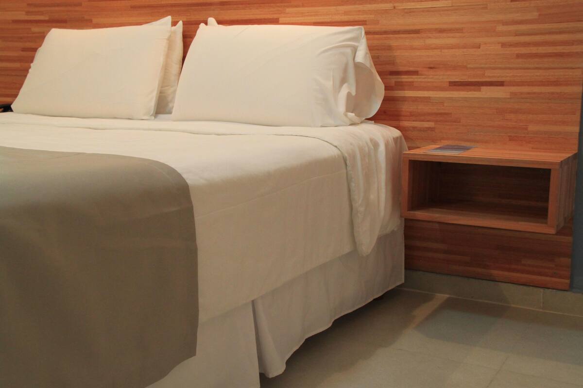 A neatly made bed with white linens and pillows, a wooden headboard, and a small wooden nightstand with a book.