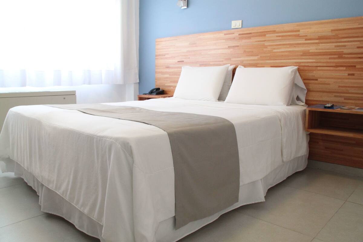 A neatly made bed with a wooden headboard, a bedside table with a lamp, and a window with sheer curtains.