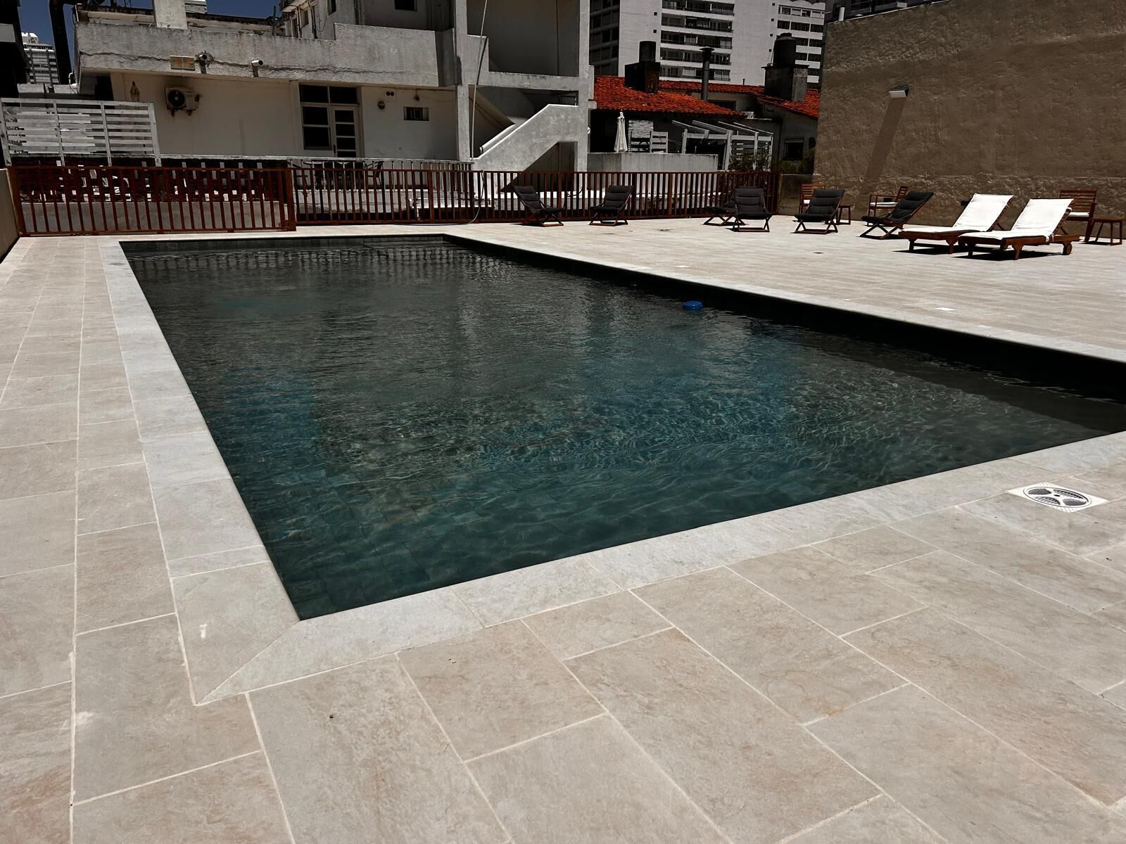 A rectangular swimming pool with clear water, surrounded by a tiled deck. There are lounge chairs and a small umbrella on the deck.