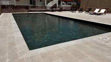 A rectangular swimming pool with clear water, surrounded by a tiled deck. There are lounge chairs and a small umbrella on the deck.