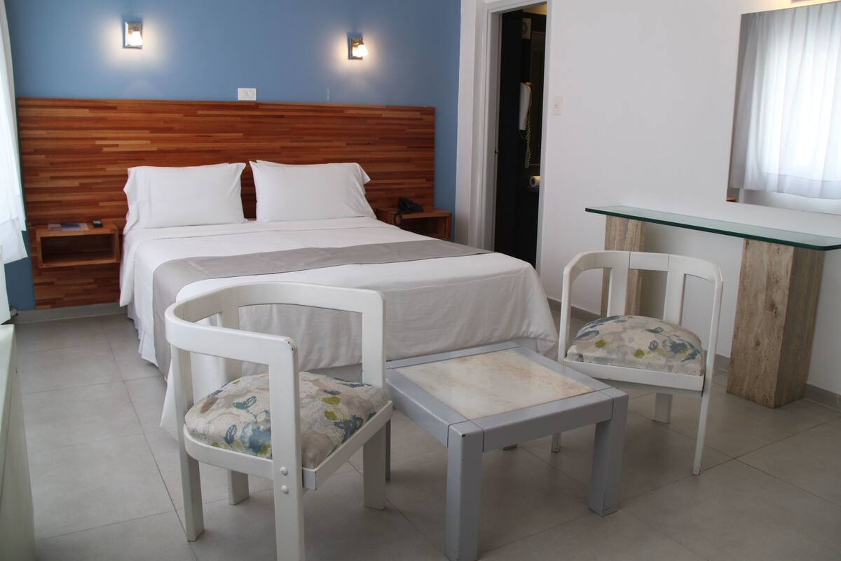 A hotel room with a wooden headboard, two beds, a small table, two chairs, and a glass desk.