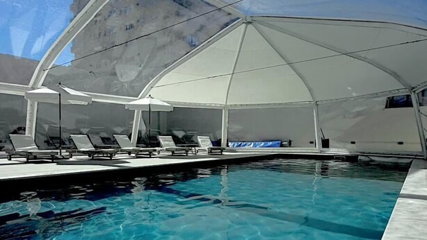 A swimming pool with a clear blue water surface, surrounded by a covered area with lounge chairs. - Hotel Florinda (Punta del Este)