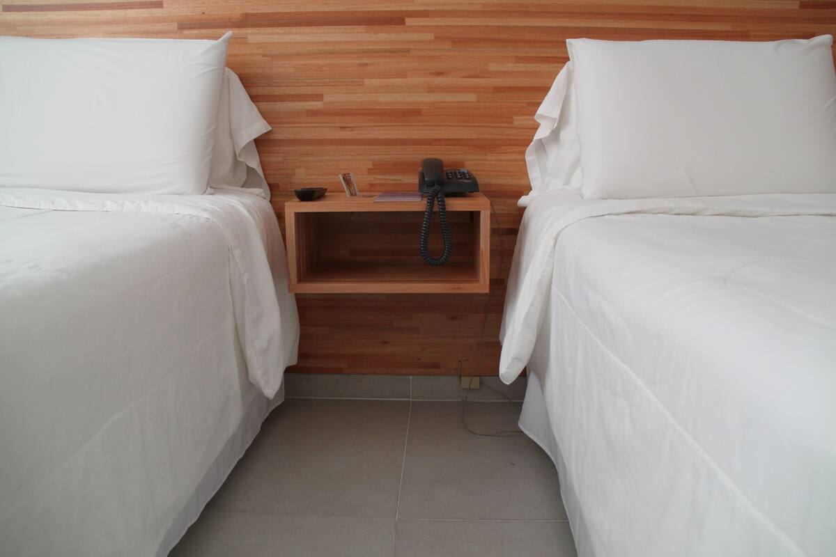 A hotel room with two single beds, a wooden nightstand with a telephone, and a small shelf with a book and a cup.