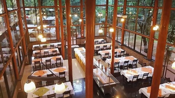 Een ruime eethoek binnen met meerdere tafels en stoelen, grote ramen met uitzicht op de groene omgeving.