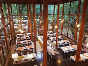 A spacious indoor dining area with multiple tables and chairs, large windows offering a view of the surrounding greenery.
