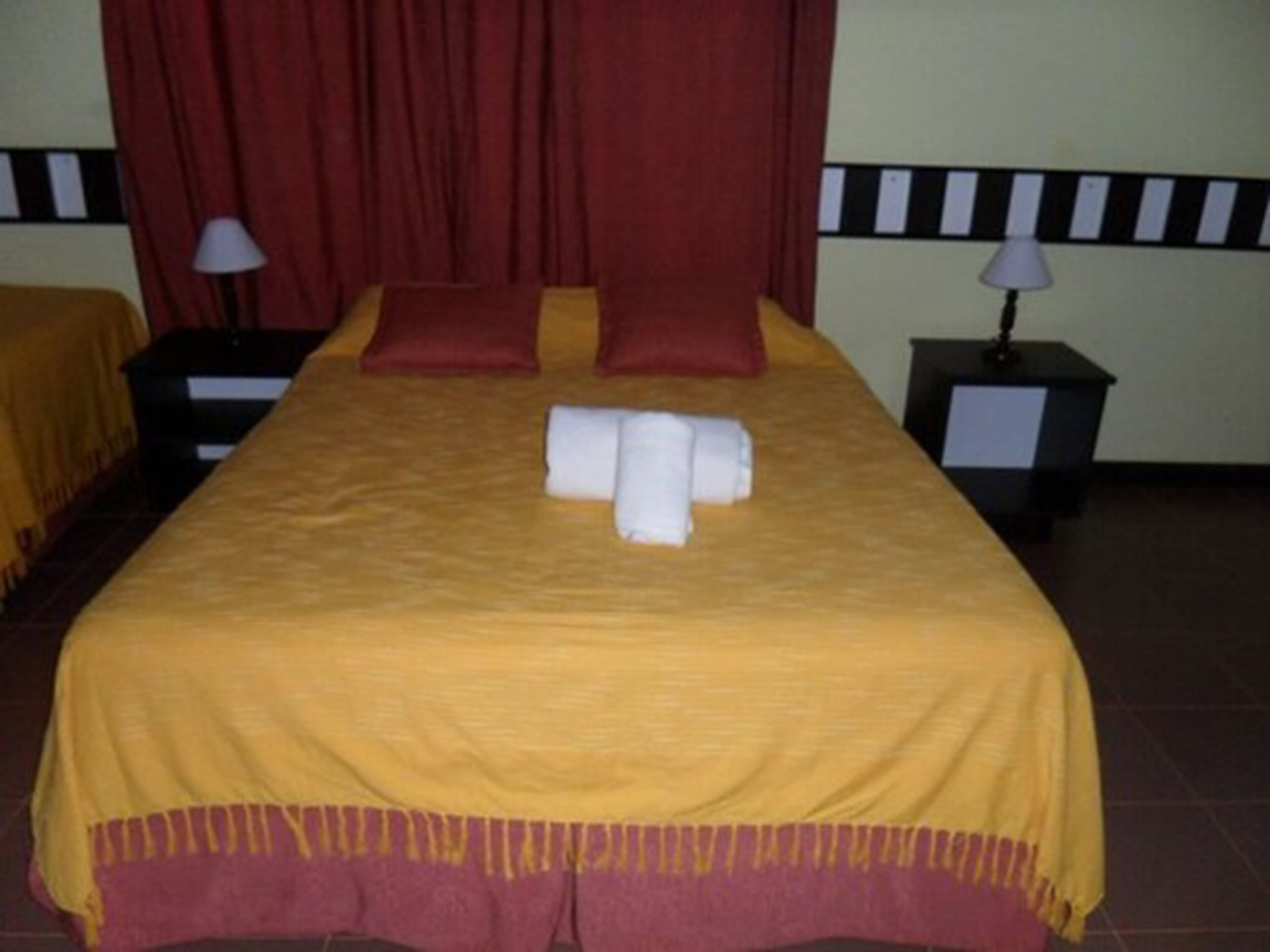 A neatly made bed with a yellow bedspread and two pillows, a pair of folded towels, and a red curtain in the background.