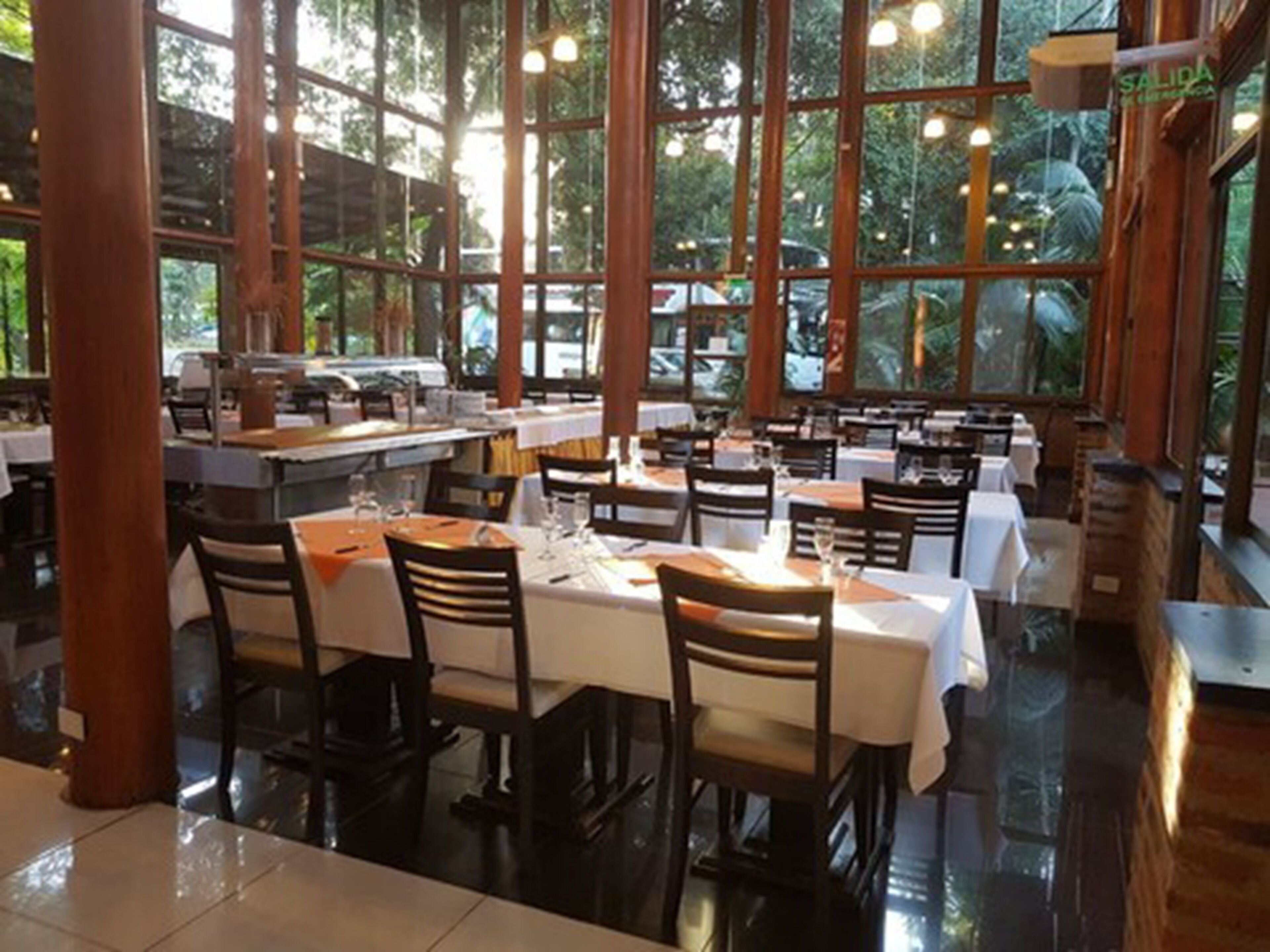 A restaurant with multiple tables and chairs, large windows, and a view of greenery outside.