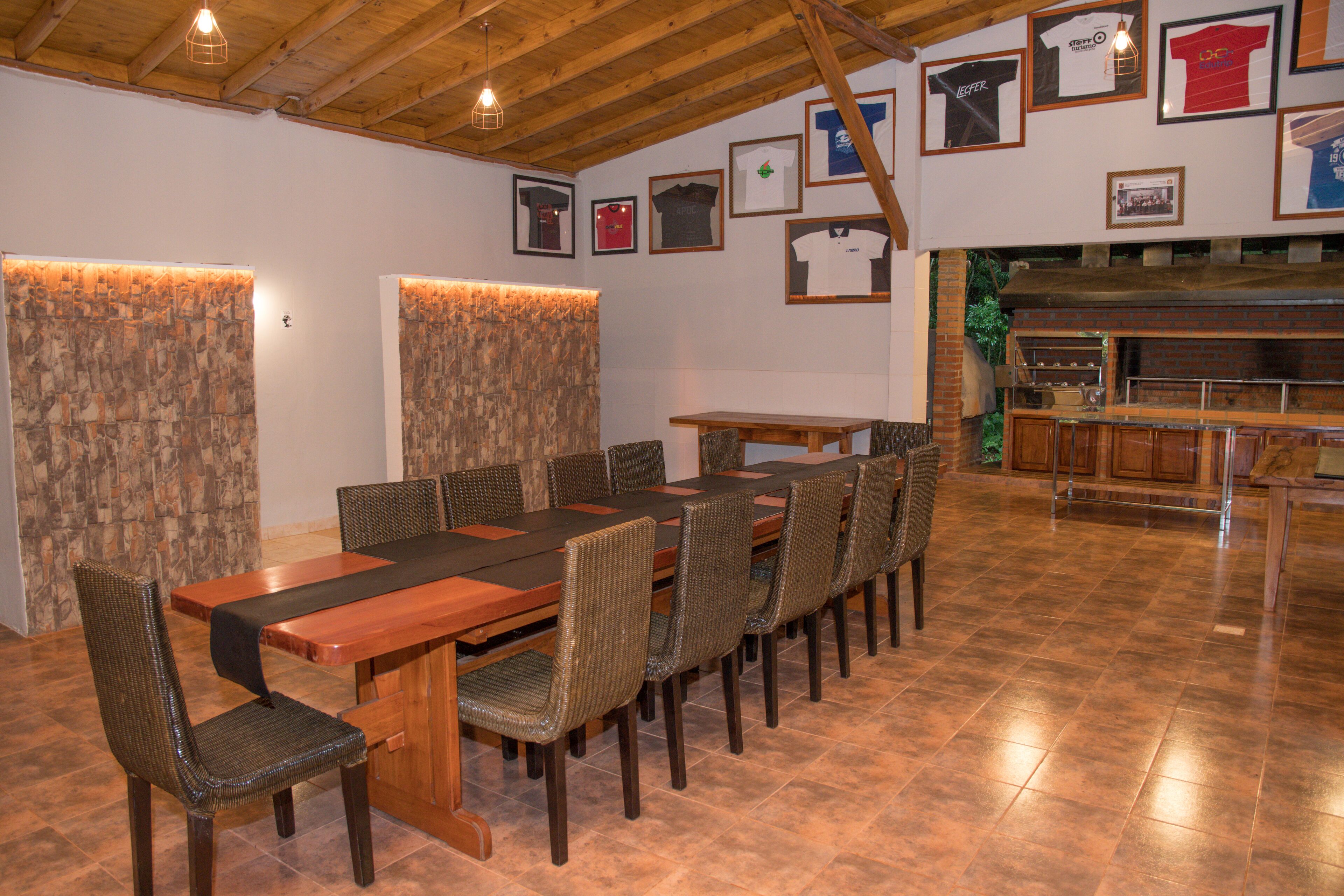 A dining area with a long wooden table and wicker chairs, a brick wall with framed sports jerseys, and a rustic wooden ceiling.