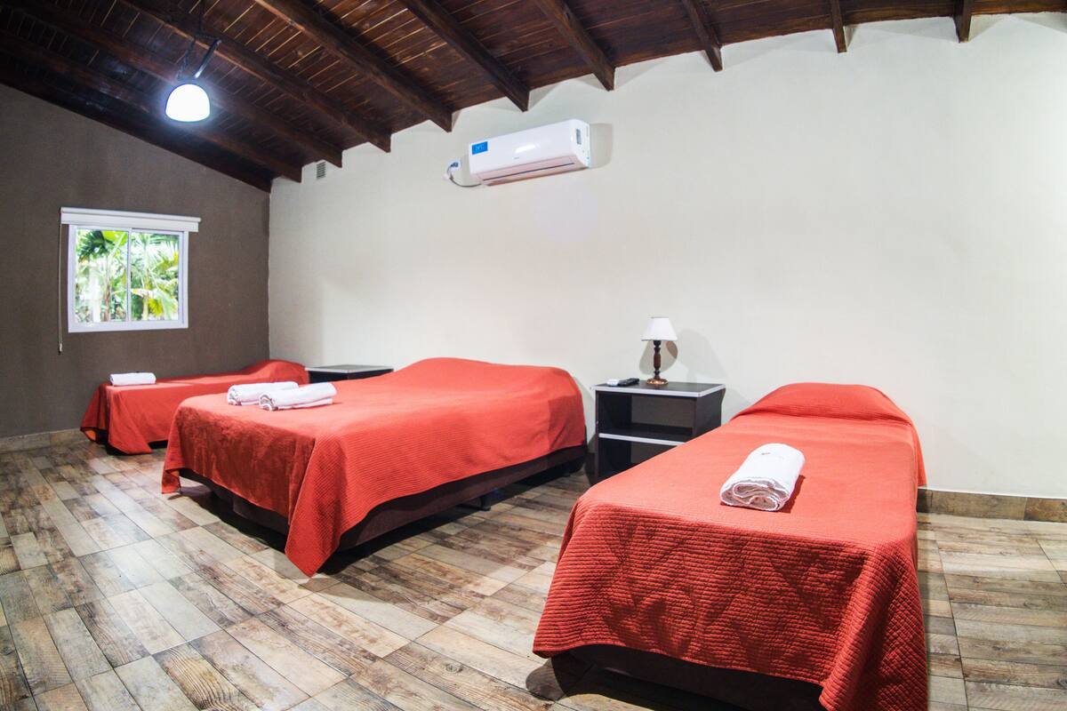 Une chambre avec trois lits, chacun recouvert d’une couverture rouge et agrémenté d’une serviette enroulée. Il y a une fenêtre donnant sur un paysage verdoyant, un climatiseur fixé au mur et une lampe sur une table de chevet.