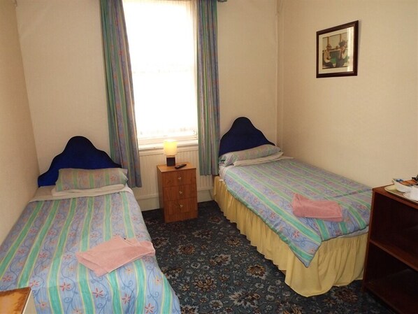 A room with two single beds, a small wooden nightstand with a lamp, a framed picture on the wall, and a desk with a chair.