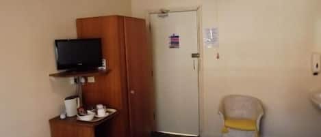 A hotel room with a bed, a television on a shelf, a chair, a small table with a cup and saucer, and a door with a sign.