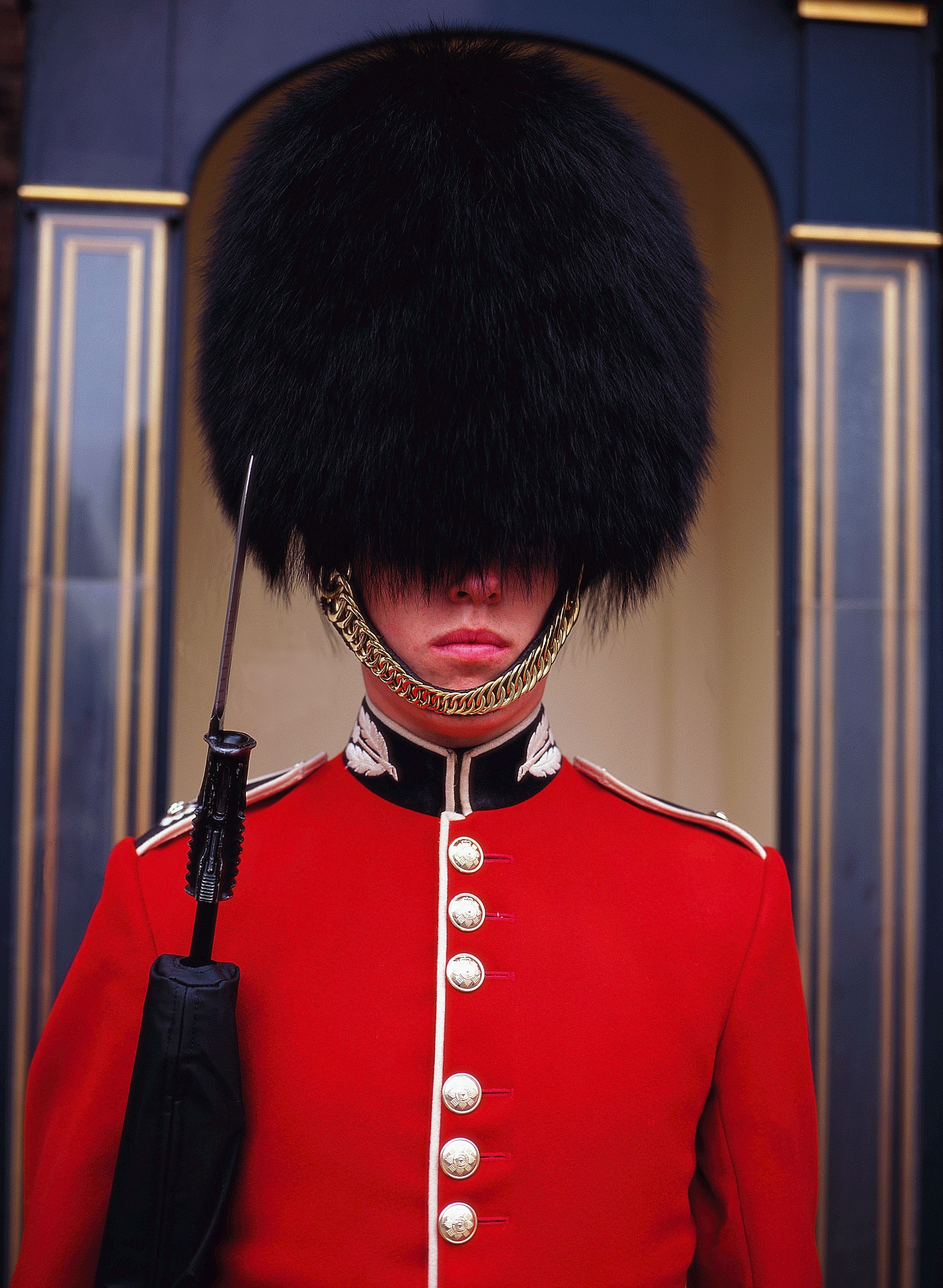 A person in a red uniform with a large black fur hat and a gold chain around the neck, standing in front of a building with a dark facade and gold trim.