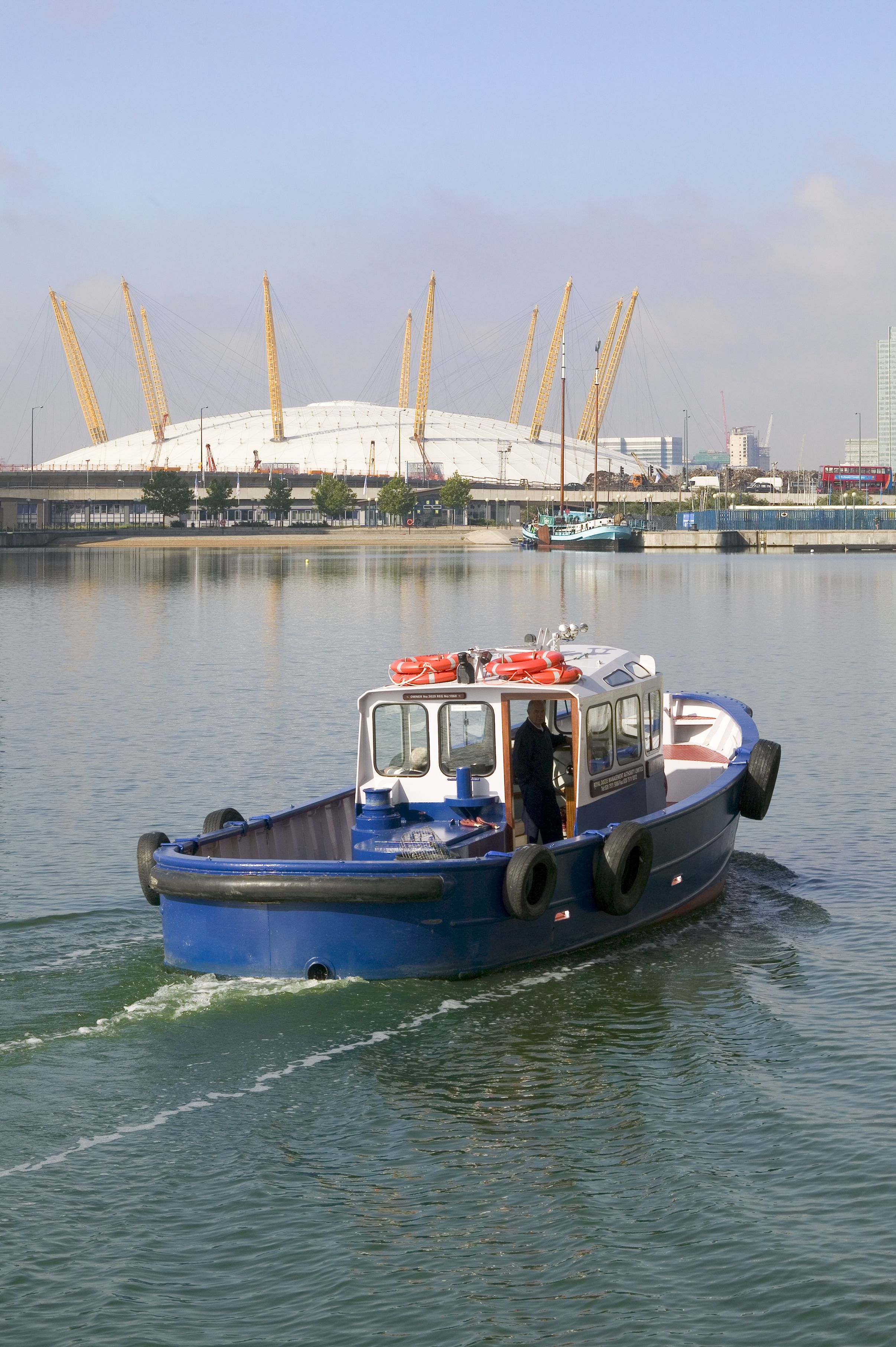 A blue boat with a cabin and lifeboats, moving on calm water with a stadium and cranes in the background.