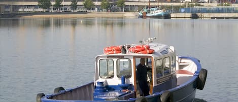 A blue boat with a cabin and lifeboats, moving on calm water with a stadium and cranes in the background.