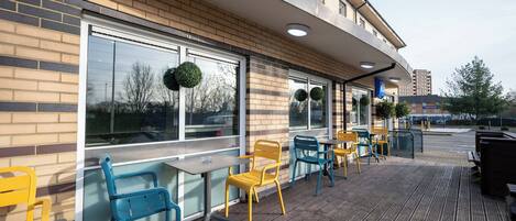 An outdoor seating area with tables and chairs, a brick wall, and large windows with wreath decorations.