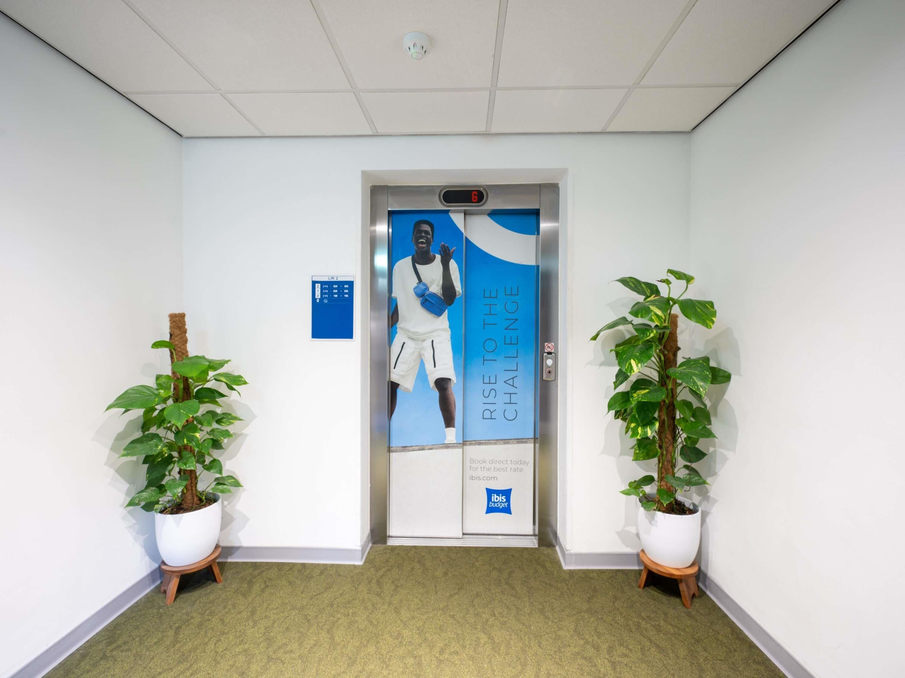 An elevator with a motivational poster, a digital display, and two potted plants.