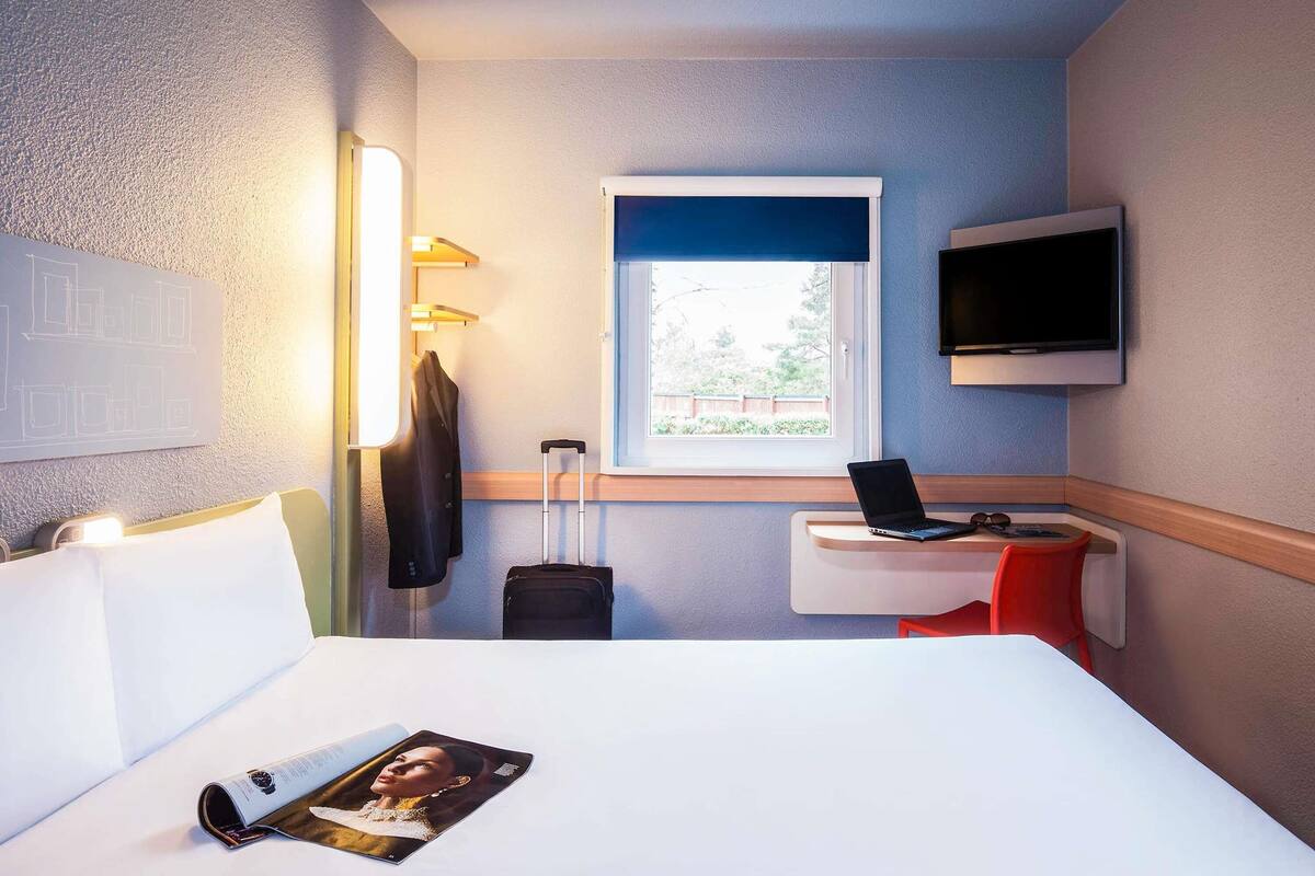 Une chambre d’hôtel avec un lit, un bureau avec un ordinateur portable, une valise, une télévision et une fenêtre donnant sur un paysage verdoyant.