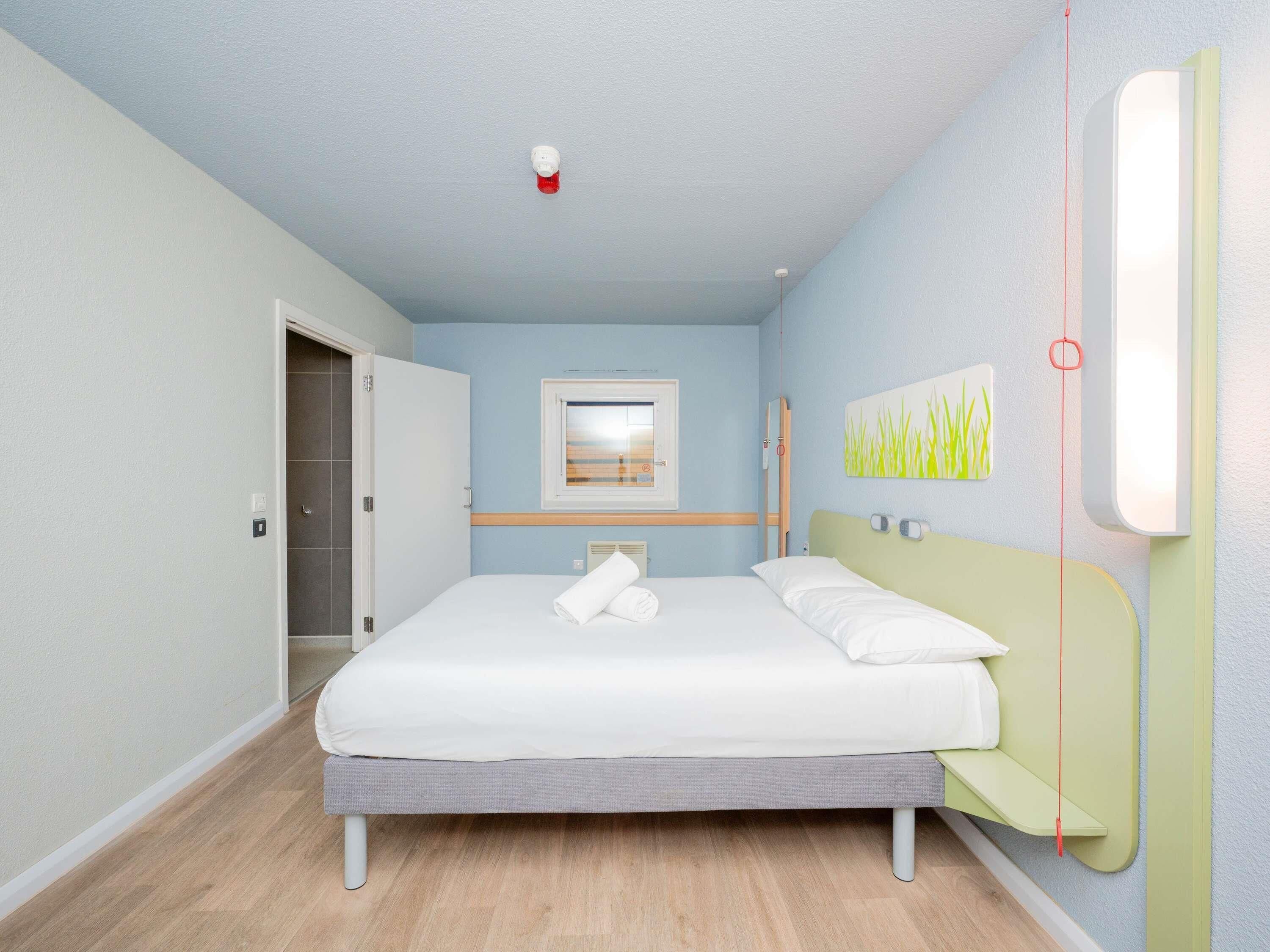 A bedroom with a bed, a window, a door, and a wall-mounted light fixture.