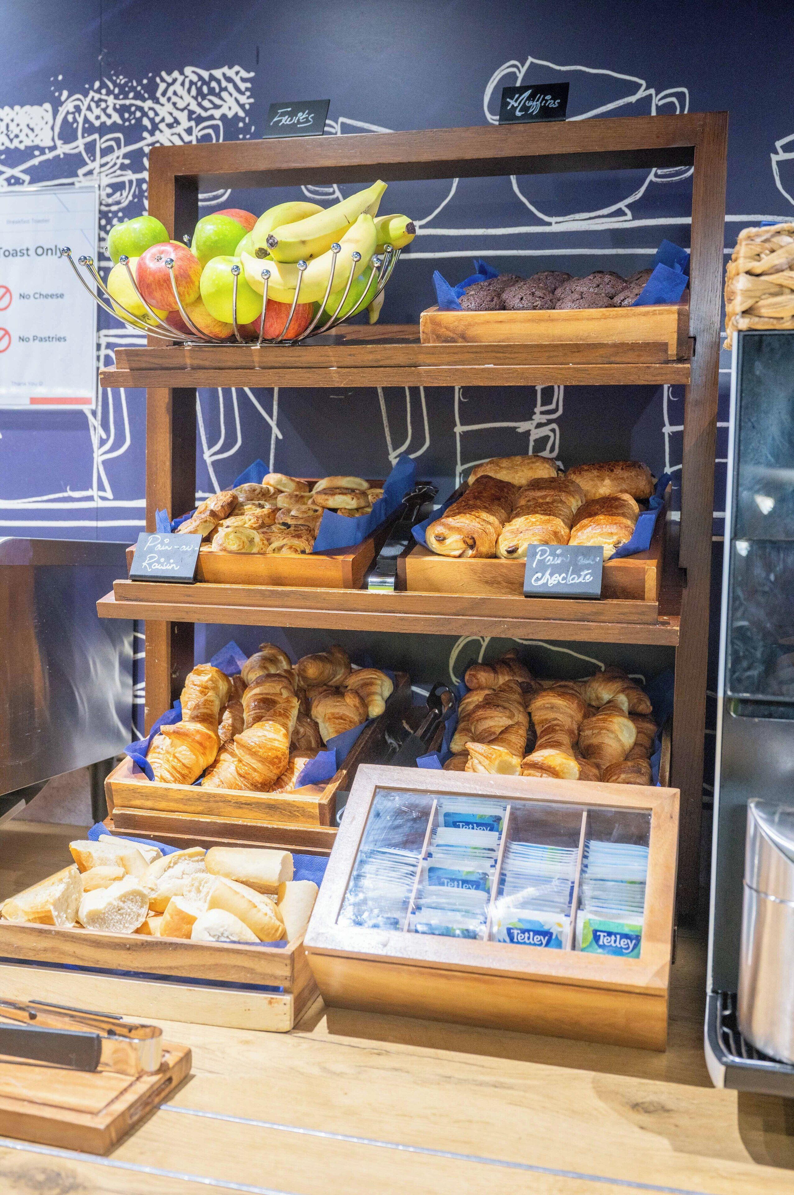 Eine Backvitrine mit verschiedenen Backwaren, darunter Äpfel, Bananen, Muffins und Gebäck.