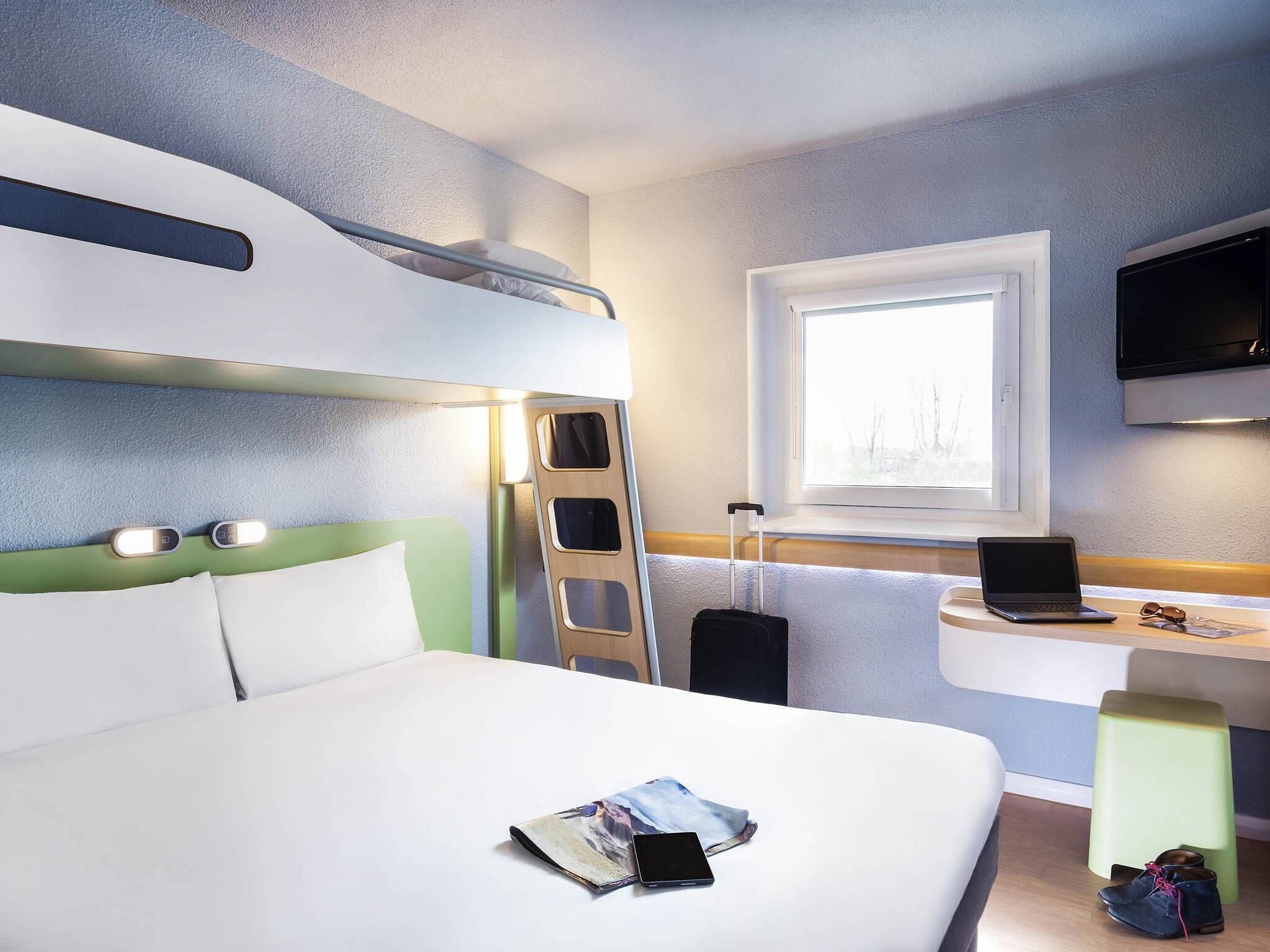 A hotel room with a bed, a desk with a laptop, a suitcase, and a TV.