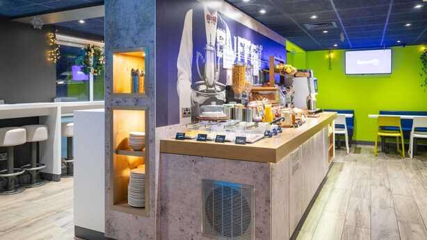 A breakfast bar with various food items and a coffee machine, surrounded by seating areas and a projection screen on the wall. - ibis budget London Barking (Barking)