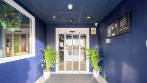 A hotel entrance with a blue wall, a glass door, and two potted plants. - ibis budget London Barking (Barking)