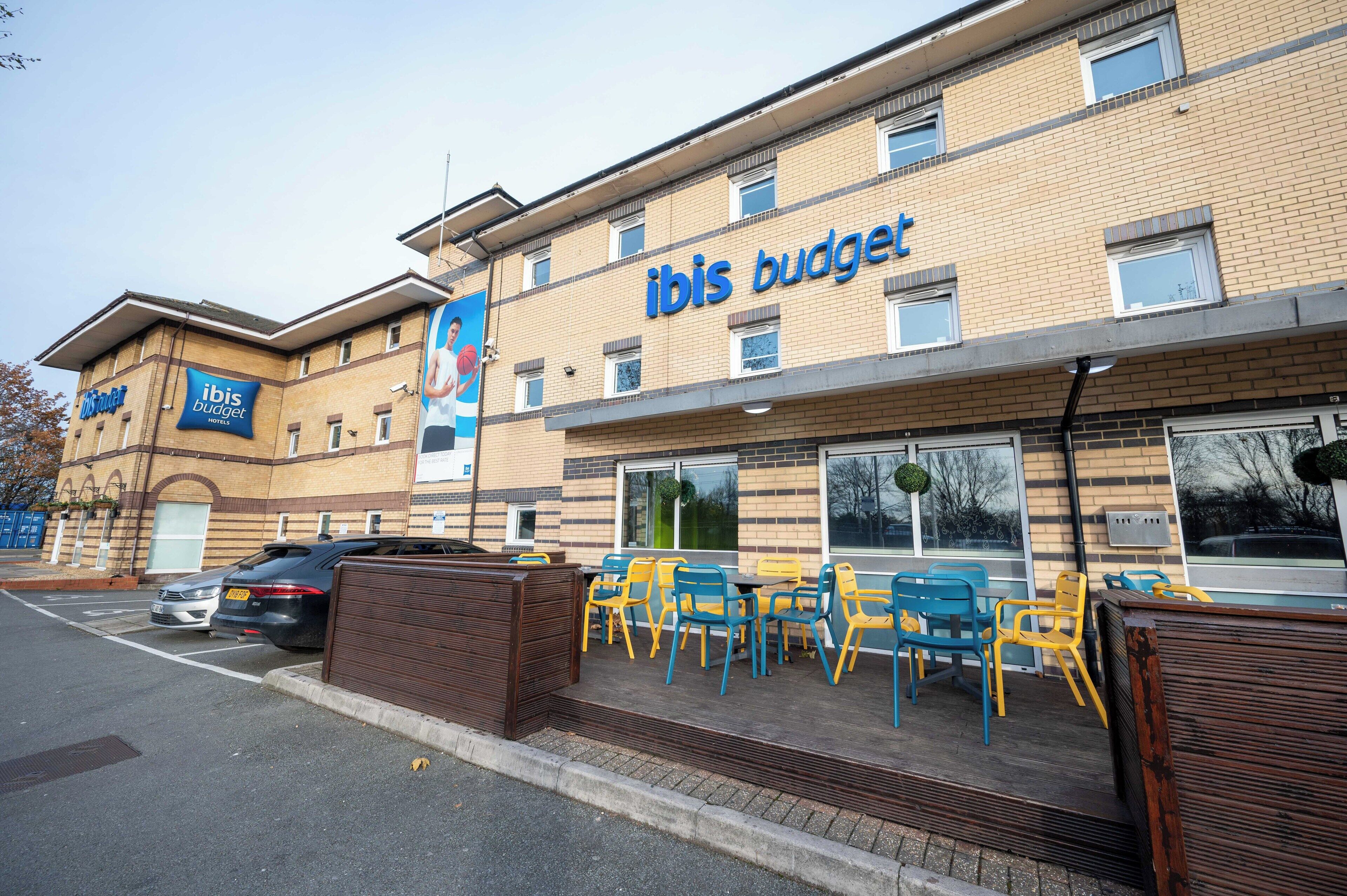 Een gebouw met twee verdiepingen met daarop het bord 'ibis budget'. Er staan buitentafels en stoelen, en een auto op de parkeerplaats.