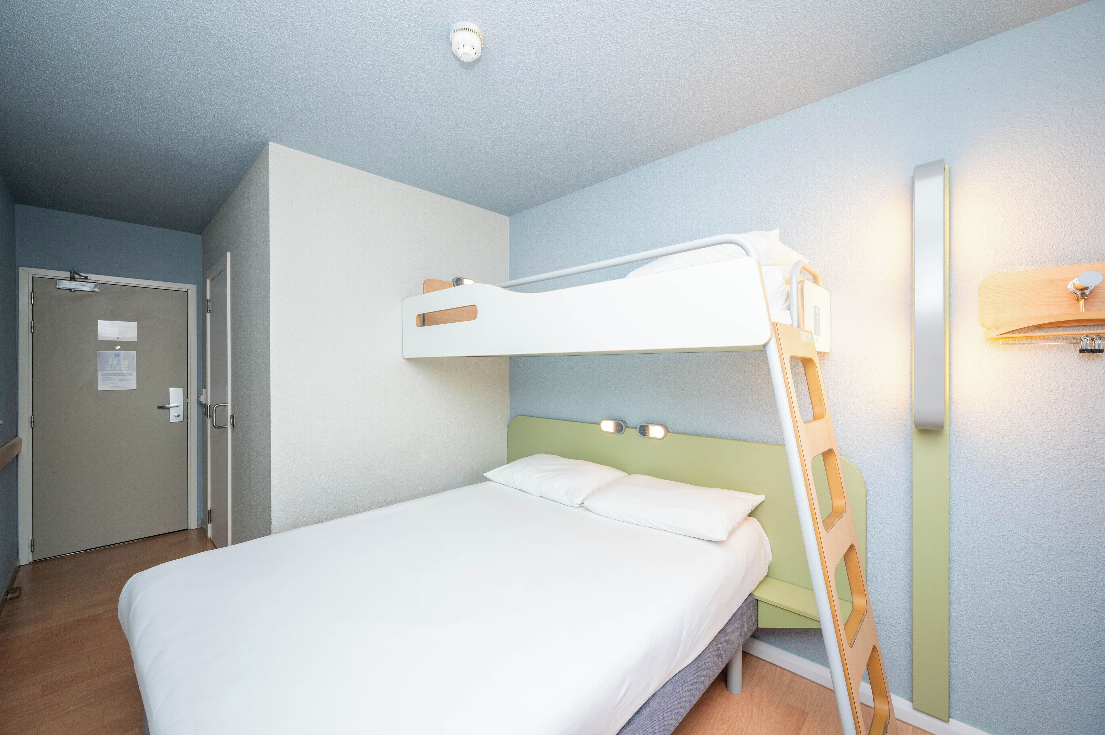 A single bed with a bunk bed above, a wooden ladder, a door with a sign, and a wall-mounted light fixture.
