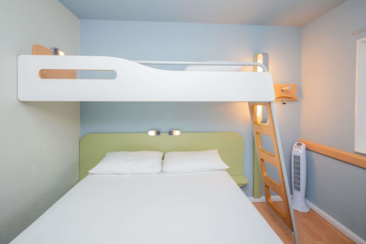 Un lit superposé avec une échelle, une table de chevet, une lampe fixée au mur et un radiateur portatif.