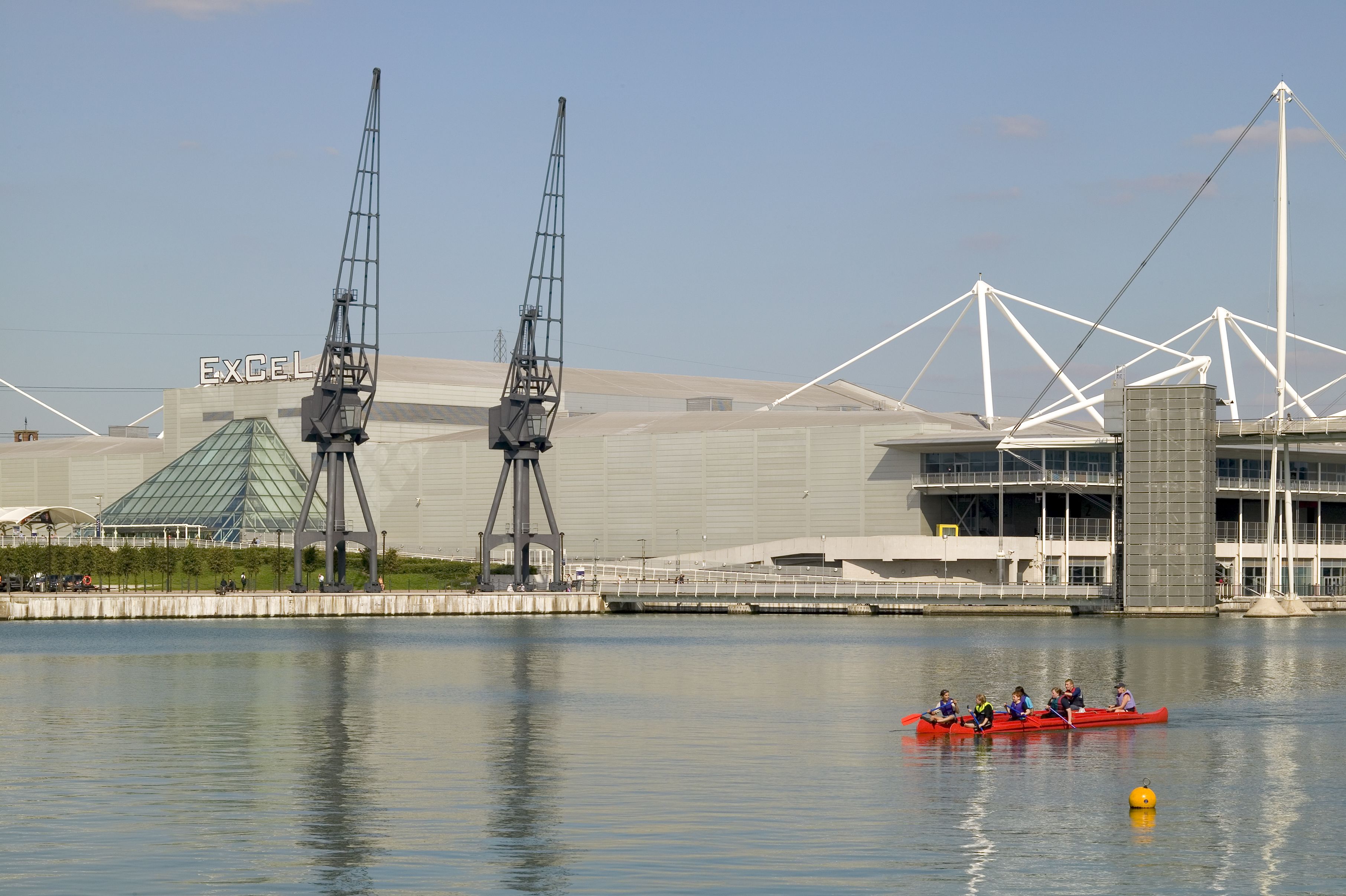 A modern building with a prominent 'EXCEL' sign, a glass pyramid, and a bridge with white trusses.