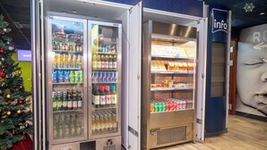 A vending machine with a variety of beverages and snacks, located in a commercial area with a Christmas tree and a wall with a large portrait. - ibis budget London Barking (Barking)
