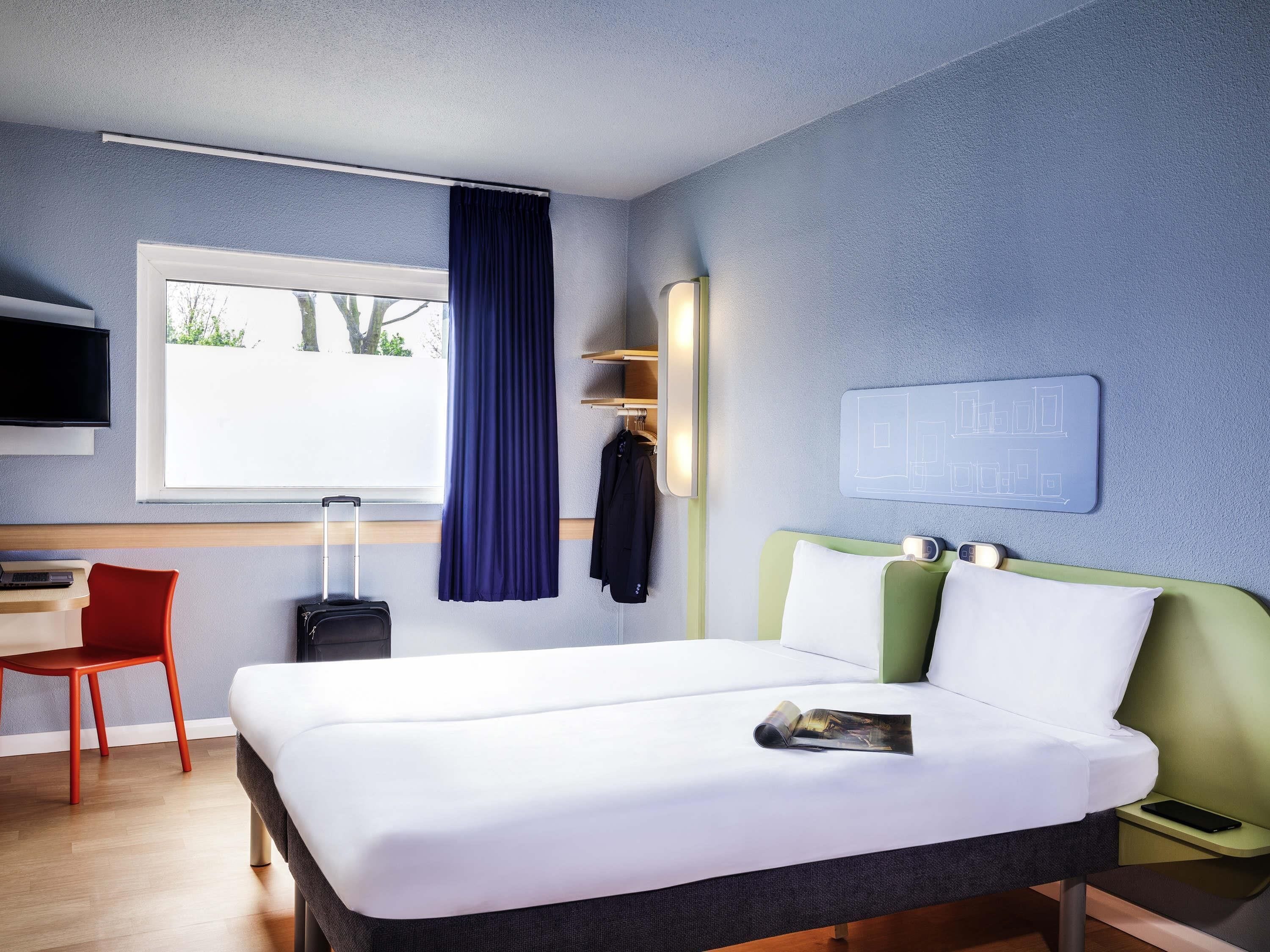 A hotel room with a bed, a desk, a chair, a TV, and a window with curtains.