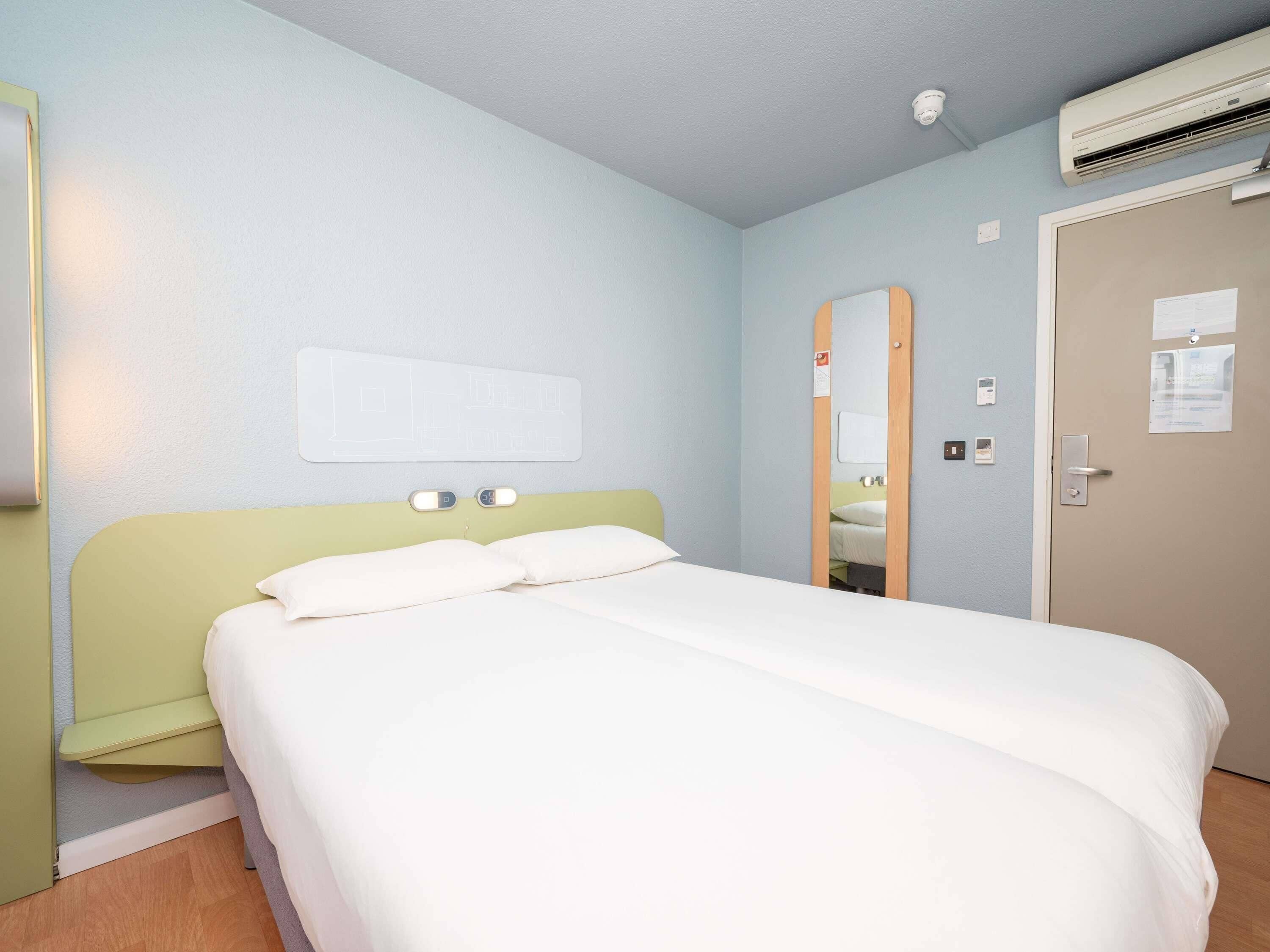 A single bed with a green headboard, a wall-mounted mirror, and an air conditioning unit above the door.