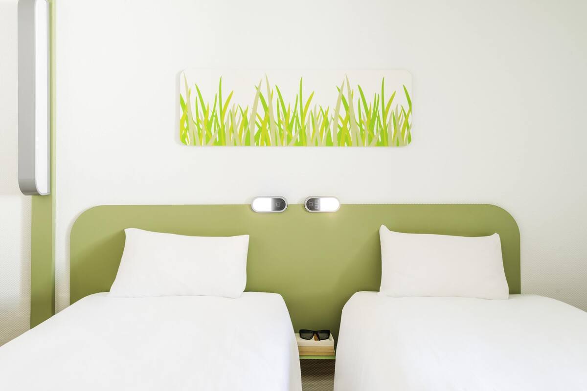 Une chambre d’hôtel avec une tête de lit verte, deux lits blancs, une lampe fixée au mur et un tableau représentant de l’herbe.