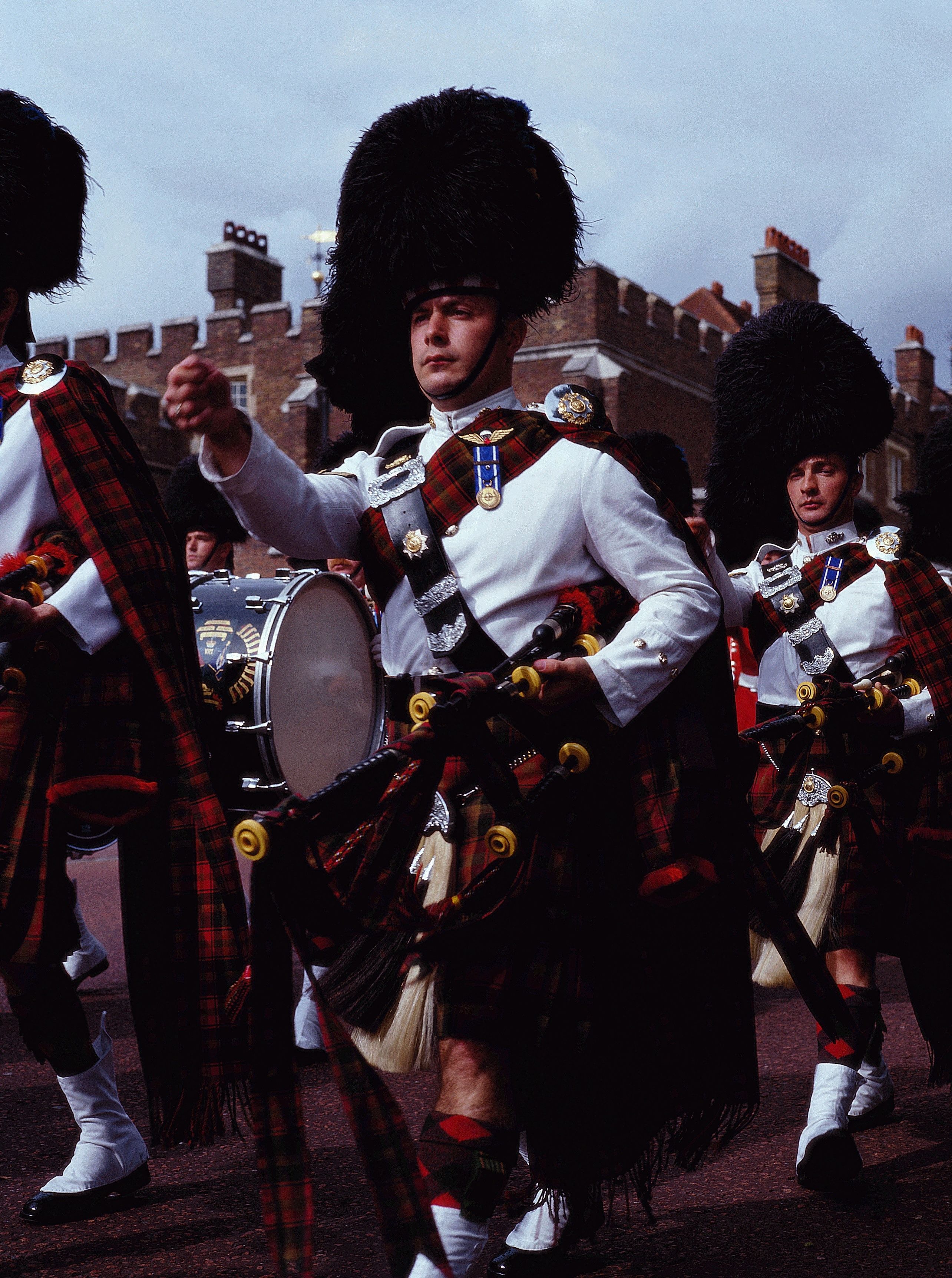 A group of individuals in traditional Scottish attire, including kilts and a large feathered hat, marching with drums.