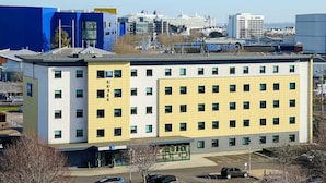 A multi-story hotel with a yellow and white facade, a parking lot with several cars, and a view of the city skyline in the background. - ibis budget Southampton Centre (Southampton)