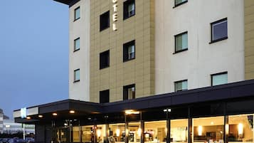 A hotel with a modern facade, large windows, and a visible restaurant area with outdoor seating.