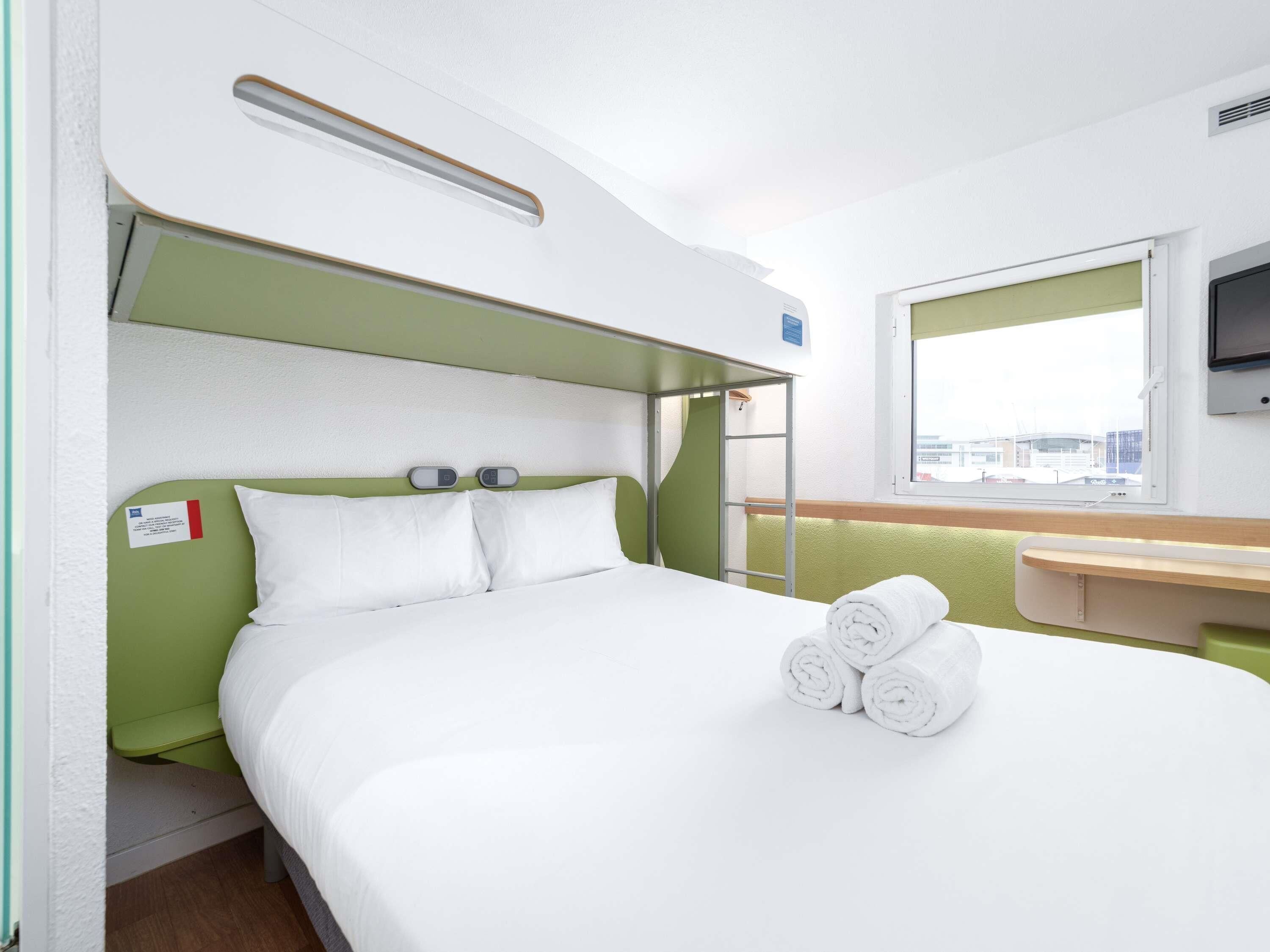 Ein Zimmer mit einem Einzelbett, einem grünen Kopfteil, einem Nachttisch, einem Fenster mit Aussicht und einem an der Wand befestigten Fernseher.