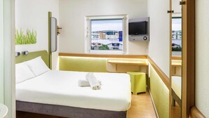 A hotel room with a bed, a desk, a TV, a window, and a view of buildings outside. - ibis budget Southampton Centre (Southampton)