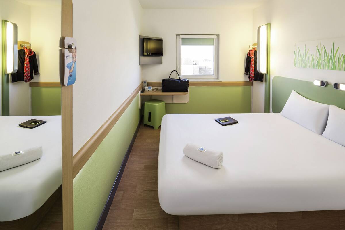 A hotel room with two beds, a TV, a small table with a handbag, and a window.