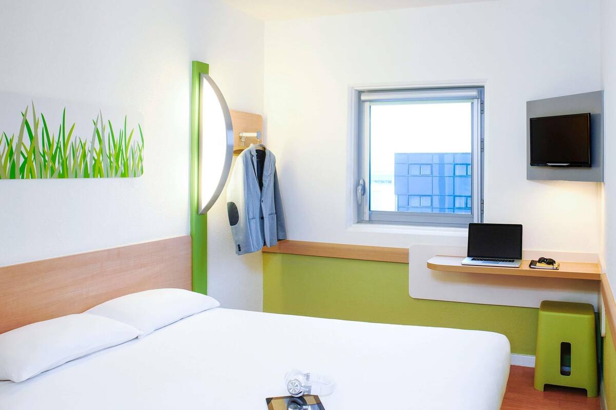 A hotel room with a bed, a desk with a laptop, a TV, and a window with a view of buildings.
